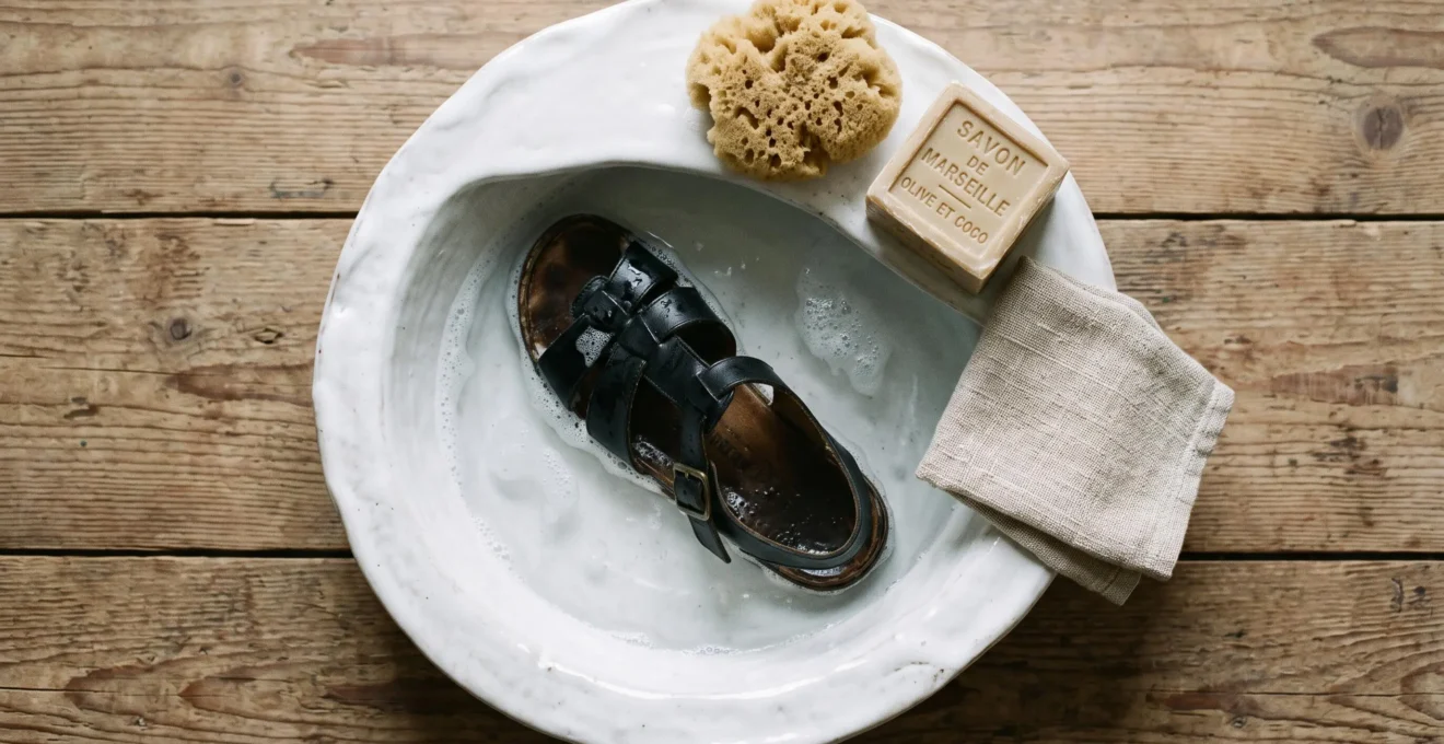 Sandale en cuir noir partiellement immergée dans une bassine d'eau savonneuse, avec une éponge naturelle et un pain de savon glycériné sur le rebord